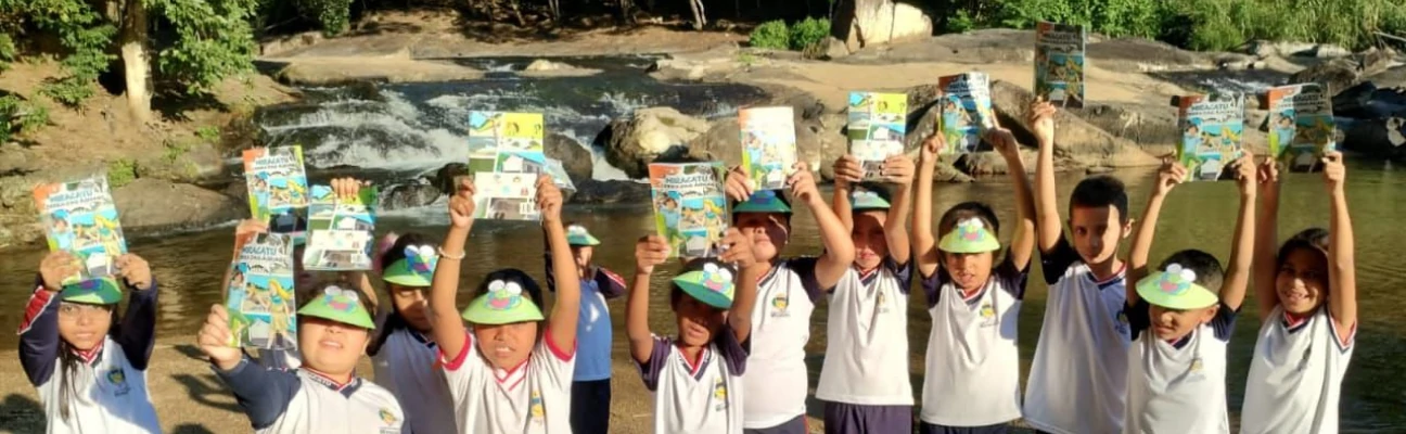 Foto de crianças com as mãos erguidas ao alto, com o livreto entre elas, e com uma cachoeira ao funto